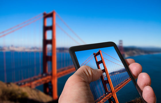 Tourist taking photo of Golden Gate Bridge in San Francisco, California on mobile gadget from top of a hill. Hand holding smartphone. Travel concept