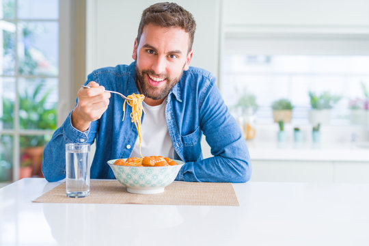 Handsome man eating pasta with meatballs and tomato sauce at home while smiling at the camera