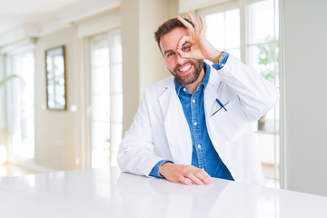 Fototapeta premium Handsome doctor man wearing medical coat at the clinic doing ok gesture with hand smiling, eye looking through fingers with happy face.