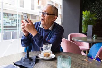 Handsome middle age senior man drinking coffee at restaurante, smiling happy enjoying and relaxing retirement using smartphone