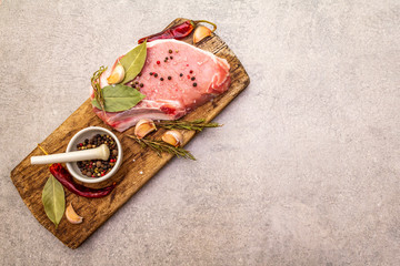 Raw pork steak with spices and dried herbs on vintage wooden board. Salt, garlic, hot pepper, rosemary, bay leaf with ceramic mortar and pestle on a stone background, top view.