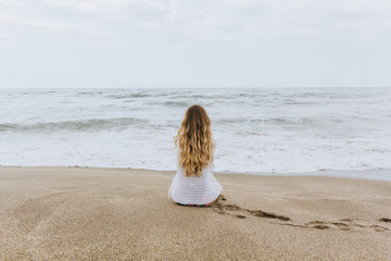 beautiful young girl woman sits on the beach, dreaming and looking at the sea. space for text. sunlight. A look from the back, against the background of sea and ocean waves