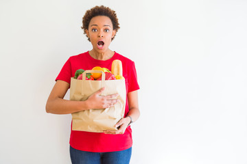 Young african american woman holding paper bag full of fresh groceries scared in shock with a surprise face, afraid and excited with fear expression