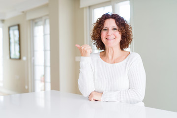 Beautiful senior woman wearing white sweater at home smiling with happy face looking and pointing to the side with thumb up.