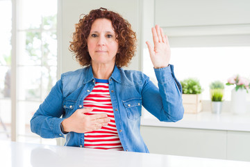 Middle age senior woman with curly hair wearing denim jacket at home Swearing with hand on chest...