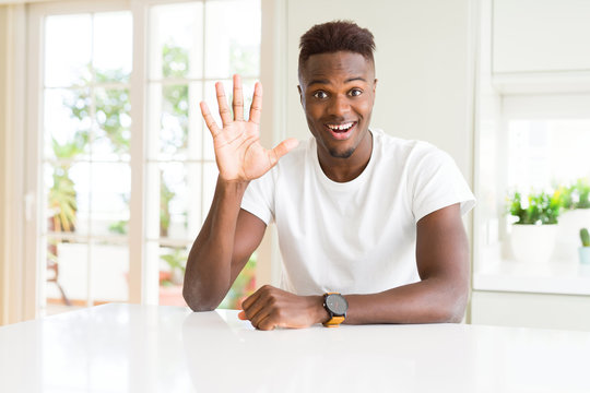 Handsome african american man on white table at home showing and pointing up with fingers number five while smiling confident and happy.