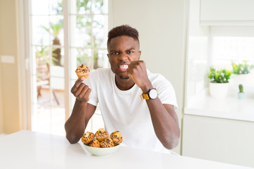African american man eating chocolate chips muffin annoyed and frustrated shouting with anger, crazy and yelling with raised hand, anger concept