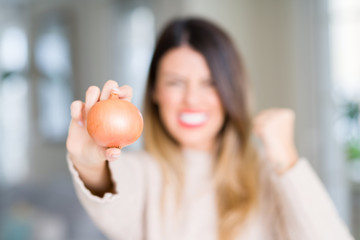 Young beautiful woman holding fresh onion at home annoyed and frustrated shouting with anger, crazy and yelling with raised hand, anger concept