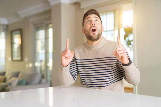 Young handsome man at home amazed and surprised looking up and pointing with fingers and raised arms.