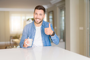 Young handsome man drinking a cup of coffee at home happy with big smile doing ok sign, thumb up with fingers, excellent sign