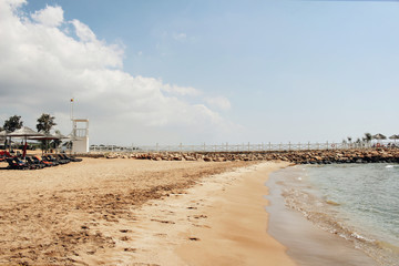 Golden sandy beach with sun beds, azure sea water and white mole in sunny day. Beautiful...
