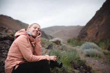 a blonde girl in a beige windbreaker sits on the rocks among the red mountains