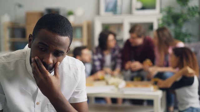 Sad African American guy with stressed face is sitting alone while cheerful friends are having fun at party eating drinking and laughing. Loneliness and problems concept.