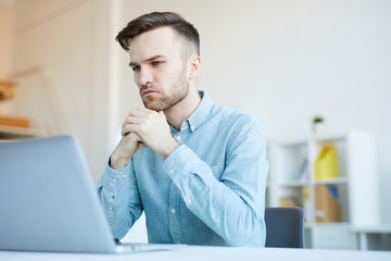 Portrait of handsome young man looking at screen thoughtfully while working in office, copy space