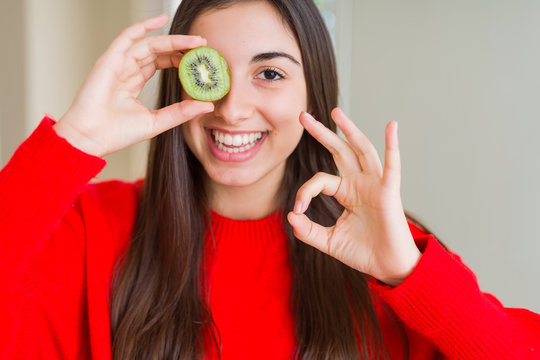 Beautiful young woman eating half fresh green kiwi doing ok sign with fingers, excellent symbol