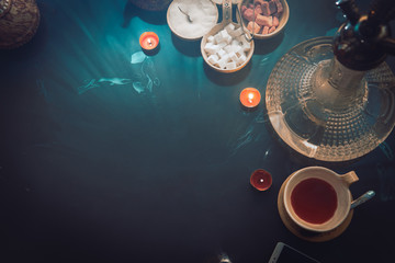 Hookah on glass table top view. cups candle and smartphone eastern tea ceremony. Stylish oriental shisha in dark with backlight. Selective focus, sugar cubes with different flavors. Copy space