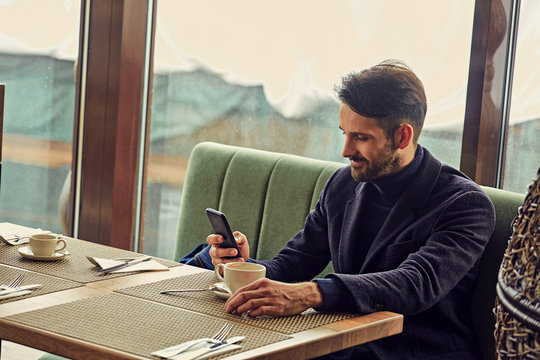Thinking handsome man sitting in cafe drinking coffee and looking in mobile phote holding in the hand. Closeup