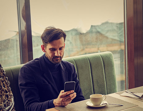 Thinking handsome man sitting in cafe drinking coffee and looking in mobile phote holding in the hand. Closeup