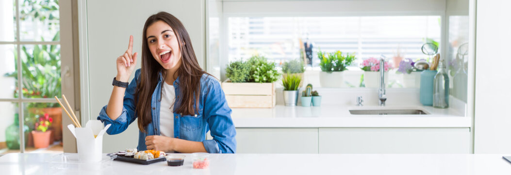 Wide angle picture of beautiful young woman eating delivery sushi pointing finger up with successful idea. Exited and happy. Number one.