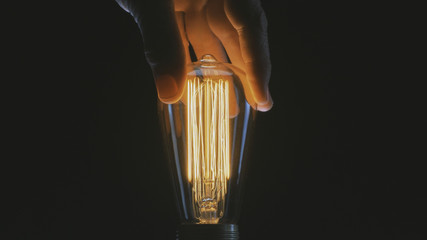 Close up shot of a male hand touching a warm vintage Edison light bulb with its glowing golden carbon thread filaments.