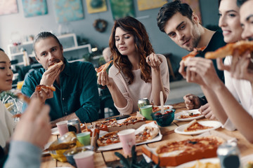 Sharing delicious meal. Group of young people in casual wear eating pizza and smiling while having a dinner party indoors