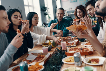 So delicious! Group of young people in casual wear eating pizza and smiling while having a dinner party indoors