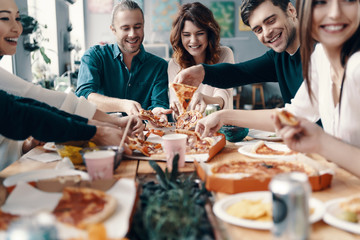 More pizza! Group of young people in casual wear picking pizza and smiling while having a dinner party indoors