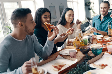 Dinner with friends. Group of young people in casual wear eating pizza and smiling while having a dinner party indoors