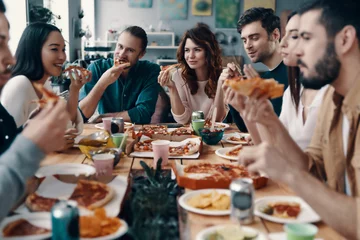 Poster Feeling hungry. Group of young people in casual wear eating pizza and smiling while having a dinner party indoors © gstockstudio