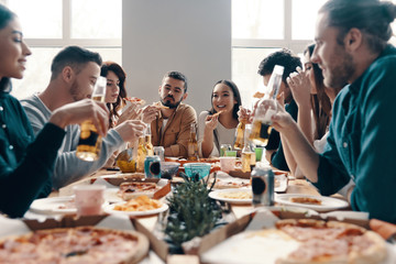 Great party. Group of young people in casual wear eating pizza and smiling while having a dinner party indoors