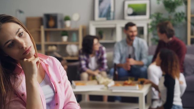 Sad girl outsider is sitting alone with unhappy face while young people are partying eating and drinking in background. Gloomy youth and celebration concept.