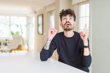 Fototapeta premium Young man wearing casual sweatshirt sitting on white table amazed and surprised looking up and pointing with fingers and raised arms.