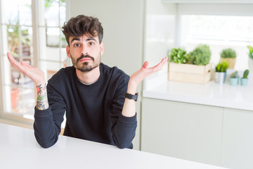 Fototapeta premium Young man wearing casual sweatshirt sitting on white table clueless and confused expression with arms and hands raised. Doubt concept.
