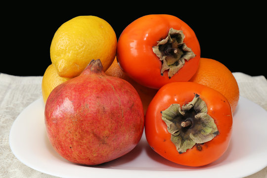 Close-up image. Still life with ripe tropical fruits. Bright orange oranges, yellow lemons, ripe and juicy persimmons and pomegranates lying on a white serving plate standing on a table with a dark ba