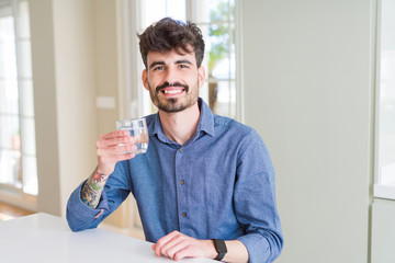 Young man drinking a fresh glass of water with a happy face standing and smiling with a confident smile showing teeth