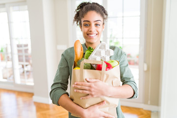 Young african american girl holding paper bag of groceries from supermarket with a happy face...