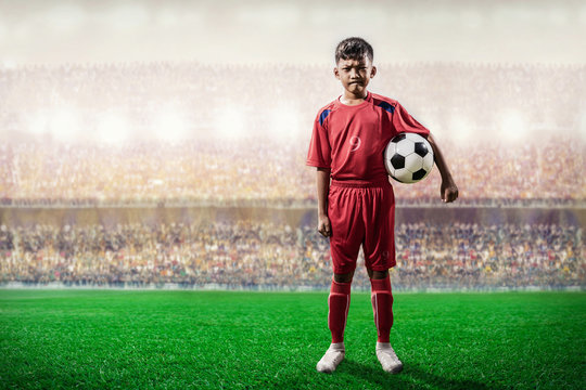 Asian Soccer Kids Player In Red Jersey. Standing And Pose To Camera In The Stadium