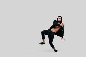 Handsome young dancer of street dancing dressed in black pants, a sweatshirt on a naked torso and a hood dances on a white background
