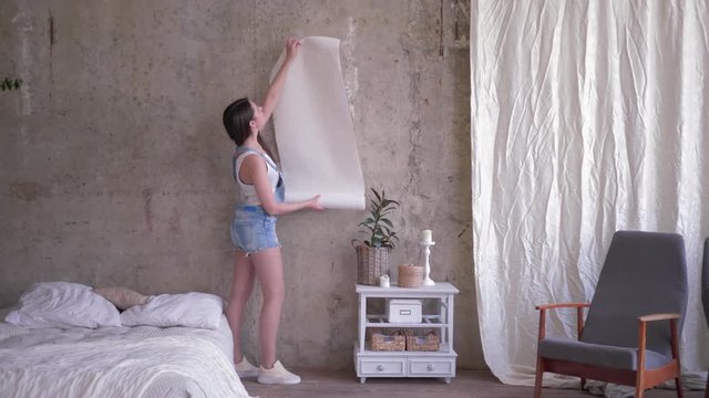 portrait of smile owner girl holding a roll of new wallpaper near the empty wall then looks on camera during renovation in room