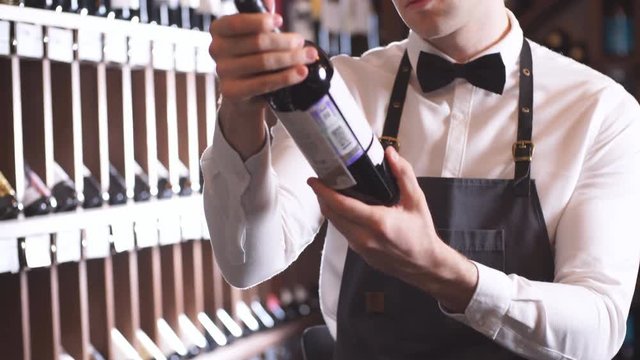 Handsome and young smiling brown haired sommelier with a bottle of wine on the background of dark wine house with shelves of bottles with alcohol