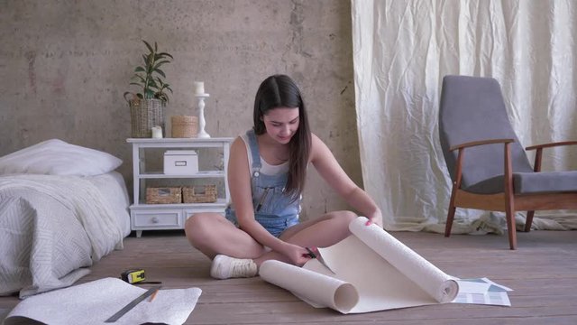 repair apartment, young woman in denim overalls cuts new wallpaper roll with scissors then view with a smile on a cut piece sitting on floor indoor