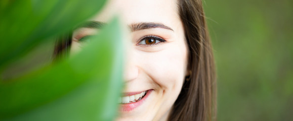 Beautiful natural young woman closeup behind big monstera leaf with green background in the woods.