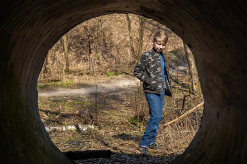 Full length portrait of one young kid standing alone in front of huge abandoned scary concrete pipe. Horizontal color photography.