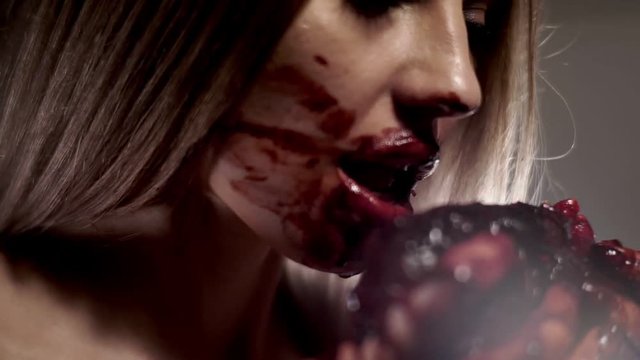 Closeup shows the face of a girl of Caucasian appearance, it is stained with blood. The girl brings the raw piece of meat and eagerly bitten him