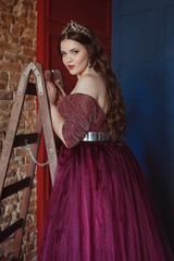 portrait of beautiful young pretty cute woman princess (queen) in long purple queen's dress and crown, with long hair and make up indoor standing on stairs on red, blue and bricks minimalistic backgro