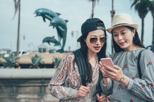 two young asian backpackers looking at pictures on mobile phone. female friends talking discussing about information on cellphone. girls standing outdoor near pool with dolphins statue under blue sky