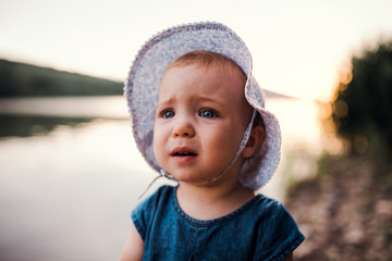 A sad small toddler girl standing outdoors by a river in summer.