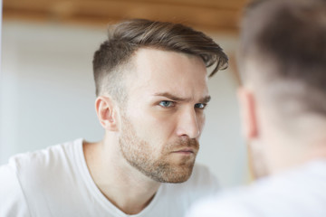 Fototapeta premium Head and shoulders portrait of handsome young looking at his reflection in mirror and frowning, copy space