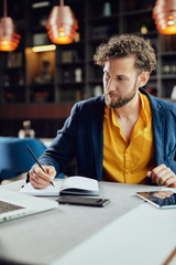 Fototapeta premium Young serious bearded Caucasian blogger dressed smart casual writing notes in agenda and looking at laptop while sitting in cafeteria.