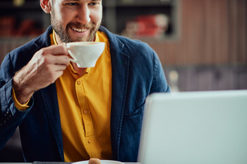 Close up of bearded smiling Caucasian freelancer sitting in cafe, drinking fresh coffee and using lapotp.